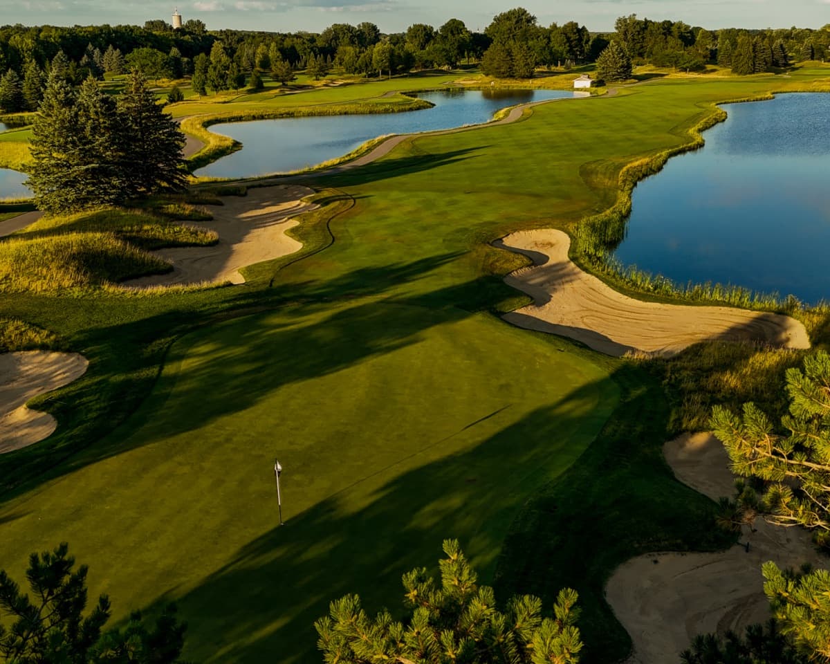 Aerial of the 13th green at The Fortress wrapped by water and fescue-edged bunkers in summer evening light